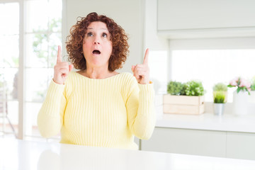 Poster - Beautiful senior woman wearing yellow sweater amazed and surprised looking up and pointing with fingers and raised arms.