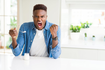 Poster - African american man eating healthy natural yogurt with a spoon annoyed and frustrated shouting with anger, crazy and yelling with raised hand, anger concept