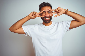 Young indian man wearing t-shirt standing over isolated white background Doing peace symbol with fingers over face, smiling cheerful showing victory