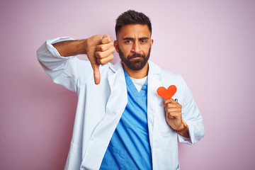 Sticker - Young indian doctor man holding paper heart standing over isolated pink background with angry face, negative sign showing dislike with thumbs down, rejection concept