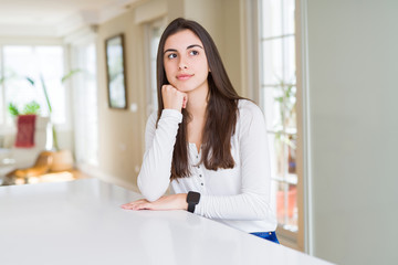 Sticker - Beautiful young woman sitting on white table at home with hand on chin thinking about question, pensive expression. Smiling with thoughtful face. Doubt concept.