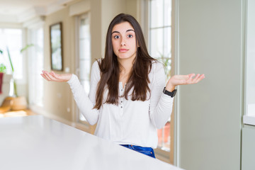 Poster - Beautiful young woman sitting on white table at home clueless and confused expression with arms and hands raised. Doubt concept.