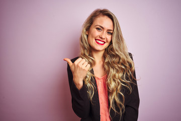 Wall Mural - Young beautiful business woman wearing elegant jacket standing over pink isolated background smiling with happy face looking and pointing to the side with thumb up.