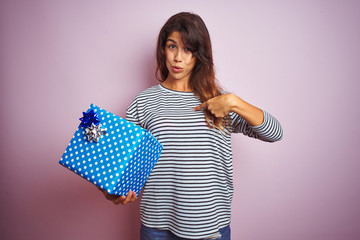 Wall Mural - Young beautiful woman holding birthday gift over pink isolated background with surprise face pointing finger to himself