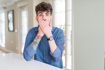 Poster - Young man wearing casual shirt sitting on white table shocked covering mouth with hands for mistake. Secret concept.