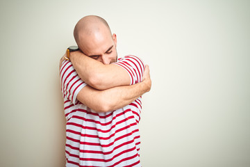 Young bald man with beard wearing casual striped red t-shirt over white isolated background Hugging oneself happy and positive, smiling confident. Self love and self care