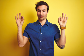 Sticker - Young handsome man with tattoo wearing casual shirt standing over isolated yellow background relax and smiling with eyes closed doing meditation gesture with fingers. Yoga concept.