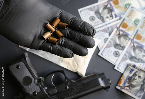 Gun lying on the table. Man in black gloves holding bullets. Illegal drug selling. Criminal problems. Dollars.