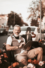 Wall Mural - Adult businessman work in cafe on the city street. Stylish bearded man write note in notebook or laptop. Male model portrait outdoors on buildings background