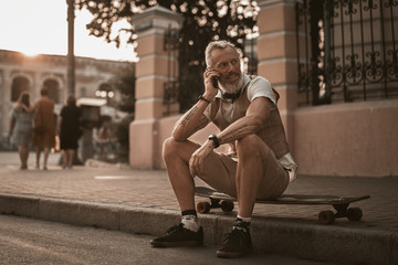 Wall Mural - Handsome adult man sitting on longboard and talk on telephone in casual clothes on city street. Stylish portrait male outdoors with skateboard.