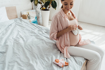 Wall Mural - smiling pregnant woman sitting on bed and holding baby shoes