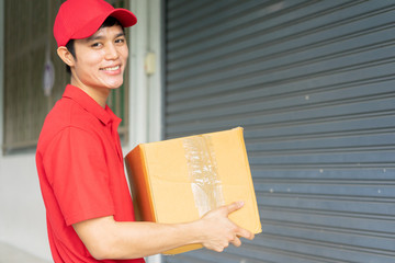 close up young postal worker (with red uniform) carrying box and stand in front of house to give order to customer for business and e-commerce lifestyle concept