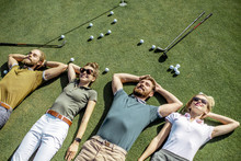 Group Of A Young And Cheerful Friends Lying On The Golf Course With Balls And Putters On The Grass, Resting And Having Fun After The Game
