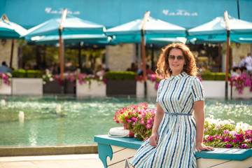 Beautiful young smiling woman in sunglasses sitting near city park fountain at summer day wearing dress and looking to the side