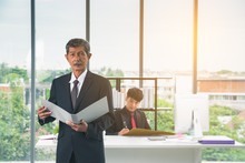 Mature Businessman Wearing Black Suit And Holding A File With A Serious Expression At Office On Soft Blurred Background Junior Businessman Sitting On Desk.