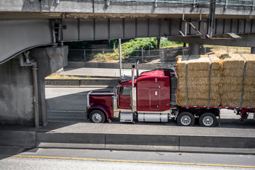 Dark red big rig classic semi Truck with flat bed semi trailer carries pressed hay running on the highway under the bridge