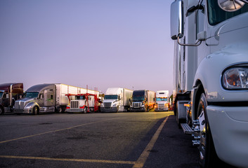 Part of big rig semi truck in front of another semi trucks standing in row on night truck stop parking lot in the headlights