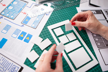 UX design. Application prototype. Phone application designer. The girl thinks what the mobile application will be. Designer desktop. Presentation of a phone app made of paper. Making app