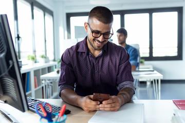 Smiling young businessman using his smartphone while working with computer in the modern startup office.