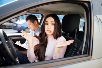 Male auto instructor takes exam in young woman. Sit beside model in car and write down. Young woman look confused on camera and show hands. Stress and failing exams.