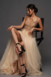 Full length fashion style portrait of young elegant woman sitting at high chair. Lady in golden evening expensive dress. Caucasian lady with hirstyle. Studio shot