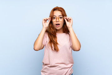 Wall Mural - Teenager redhead girl over isolated blue background with glasses and surprised