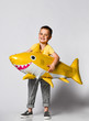 © FAB.1 - Positive little boy, dressed in festive clothes, wears a balloon in the shape of a yellow shark fish, celebrates a holiday, has a wide smile, stands on a light background, being in a good mood.