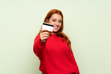 Wall Mural - Teenager redhead girl with sweater over isolated green background holding a credit card