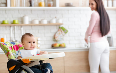 Cute baby playing on high chair in kitchen, mother washing dishes