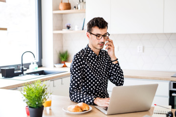 Wall Mural - Young man with laptop and smartphone sitting in kitchen, a home office concept.