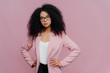© VK Studio - Photo of serious curly haired woman looks confidently at camera, wears formal neat clothes, keeps both hands on waist, wears optical glasses, poses over purple background. People, style concept