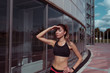 Beautiful athletic tanned girl looks into distance, covers her hand from sun, in summer city background of glass windows, free space for fitness motivation text. Sportswear top.