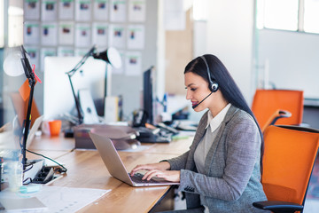 Beautiful business people in headsets are using computers and smiling while working in office. Girl is looking at camera