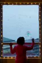 Little Girl Drawing Christmas Tree And Santa Claus On A Glass Window At Dusk
