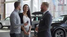 Happy Beautiful Wife And Her Cheerful Husband Speaking With Salesman And Taking Key To Their New Car While Buying In Auto Showroom