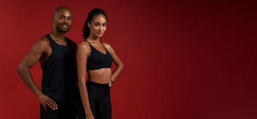 Wall Mural - Training together. Young and cheerful african fitness couple in sportswear looking at camera with smile while standing against red background