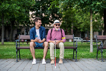 Young Man And Blind Senior With White Cane Sitting On Bench In Park In City.