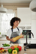 © nenadaksic - Beautiful mixed race housewife in apron standing in kitchen and holding zucchini On kitchen counter are vegetables. Meal preparation.