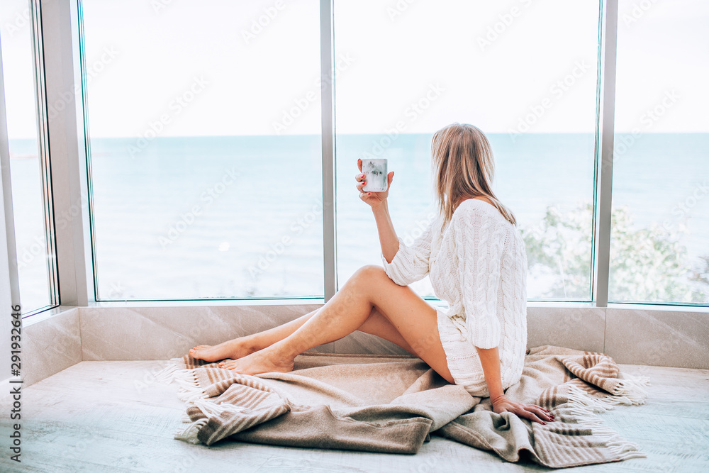 Young happy woman in a white knitted dress enjoying sea view next to big window with cup of tea. Panoramic window. Floor covering - obrazy, fototapety, plakaty 