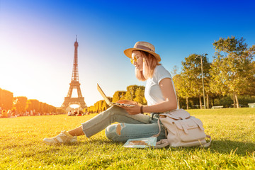 An Asian girl works on a laptop sitting on the grass on the Champ de Mars in Paris overlooking the Eiffel tower at sunset. Concept of remote work and freelancing