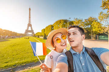 Wall Mural - Mixed race couple in love hugging and taking selfie with french flag and Eiffel tower in the background. Honeymoon travel in France and Paris concept