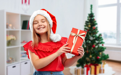 Poster - childhood and winter holidays concept - smiling happy girl in santa helper hat with gift box over christmas tree at home background