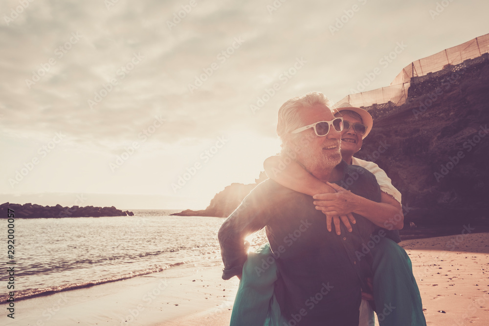 beautiful couple of seniors at the beach having fun together and playing - mature man carry his wife woman with love and forever life concept - sea or ocean in the background -   retired lifestyle - obrazy, fototapety, plakaty 