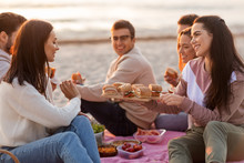 friendship, leisure and fast food concept - group of happy friends eating sandwiches or burgers at picnic on beach in summer