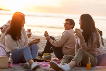 Wall Mural - friendship, leisure and fast food concept - group of happy friends eating sandwiches or burgers at picnic on beach in summer