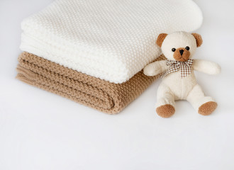 White and bronze plaids are stacked on a white surface. The teddy bear sits at them.