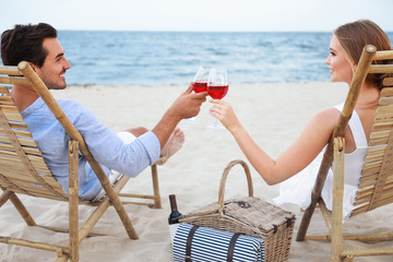 Poster - Happy young couple with glasses of wine sitting on deck chairs at sea beach