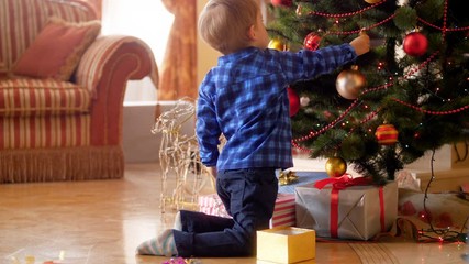 Wall Mural - 4k footage of little boy sitting on floor at living room and decorating Christmas tree with baubles and balls. Family decorating house on winter holidays and celebrations.