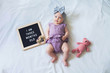 Flat lay composition of three months old baby girl laying down on white background with letter board and teddy bear.