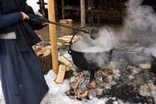 Woman In Pioneer Garb Ladling Evaporating Sap In Cast Iron Pot To Produce Maple Syrup